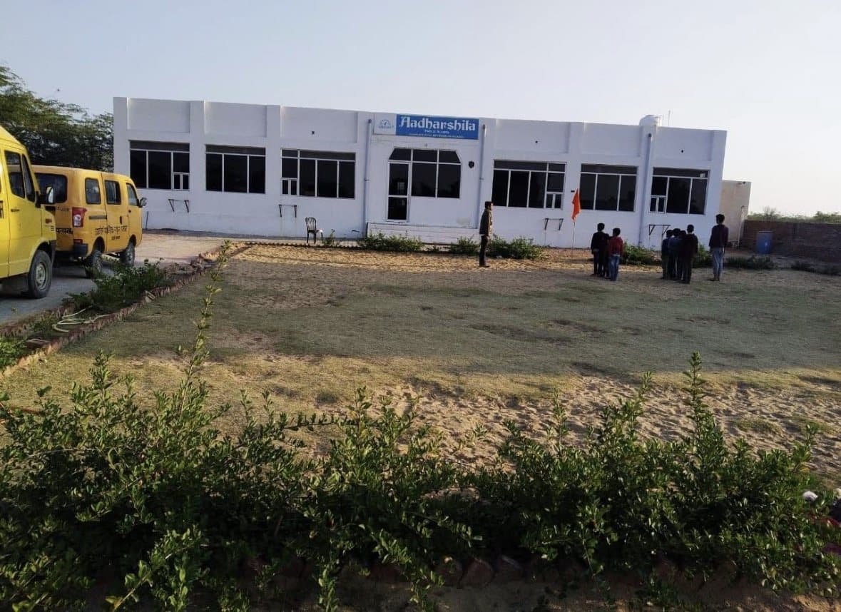 The white single-storey building of Aadharshila Public School with its blue name board, school vans parked on the left and a sandy ground edged with green shrubs.