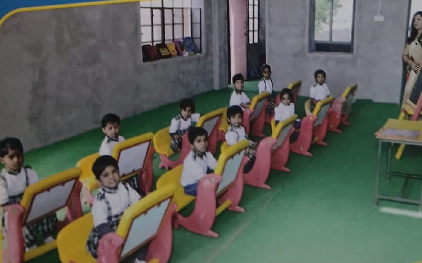 Young children seated at colourful single desks with built-in slates in a nursery classroom, a teacher standing by an easel.